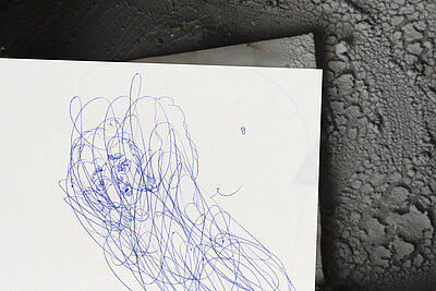 Blau ausgeführte, dichte Kritzelzeichnung eines Kopfes auf weißem Papier, das teilweise auf einer dunklen, strukturierten Oberfläche liegt. / Dense blue scribble drawing of a head on white paper, partially resting on a dark textured surface.