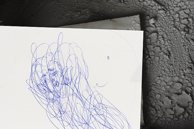 
            
                Blau ausgeführte, dichte Kritzelzeichnung eines Kopfes auf weißem Papier, das teilweise auf einer dunklen, strukturierten Oberfläche liegt. / Dense blue scribble drawing of a head on white paper, partially resting on a dark textured surface.
            
        
