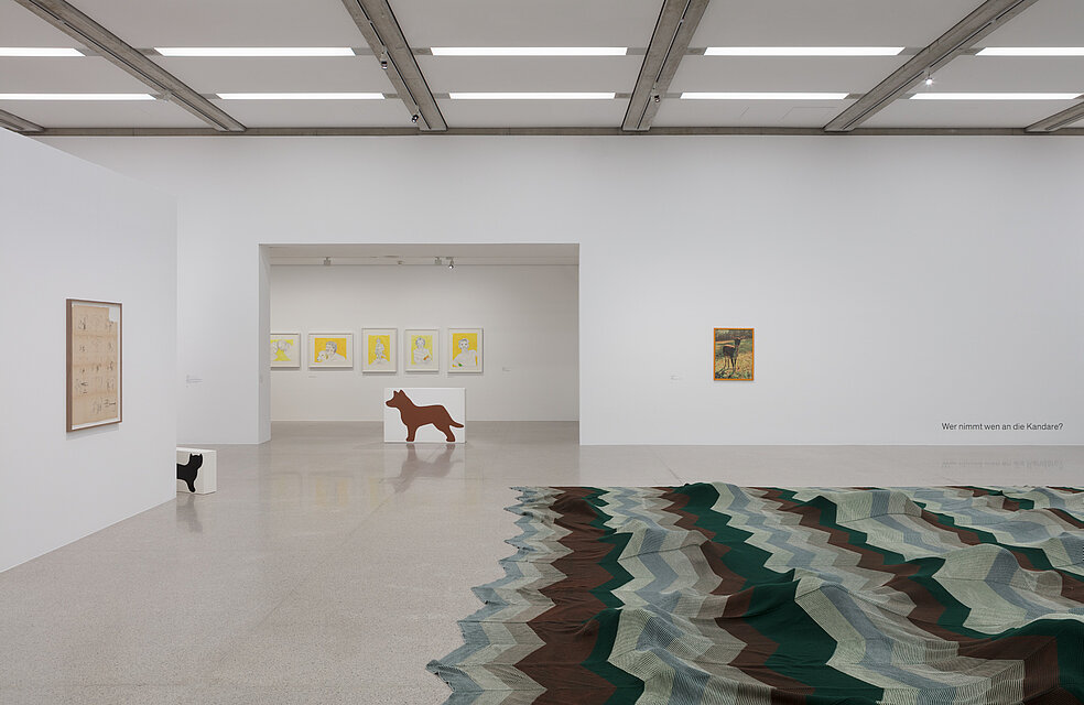 On the floor is a large-format blanket with a blue and green wave pattern. Behind it, five portraits with a yellow background hang on the wall. To the right is a picture of a deer. On the floor are two sculptures in the form of white rectangular boxes with simplified depictions of two animals. 