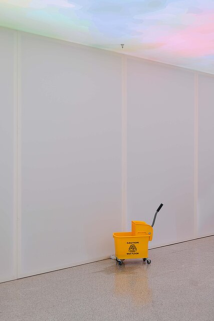 A yellow cleaning bucket stands in front of an empty wall.